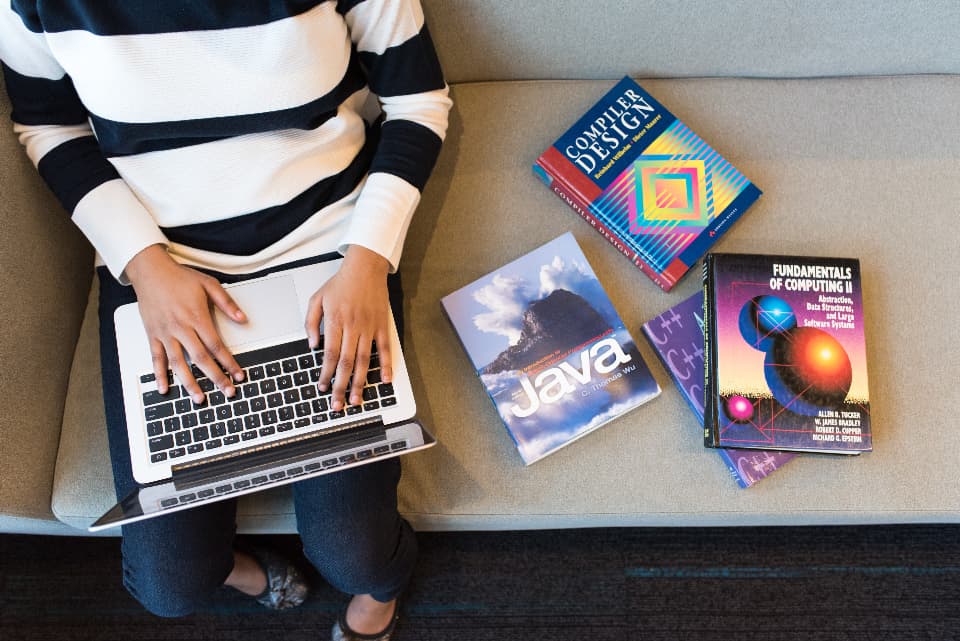 A learner working on a laptop beside a stack of computing textbooks